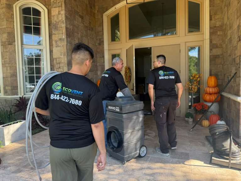 Three Ecovent technicians in black company shirts bring a NIKRO HEPA negative-air machine and its hose to the front door of a stone-faced home