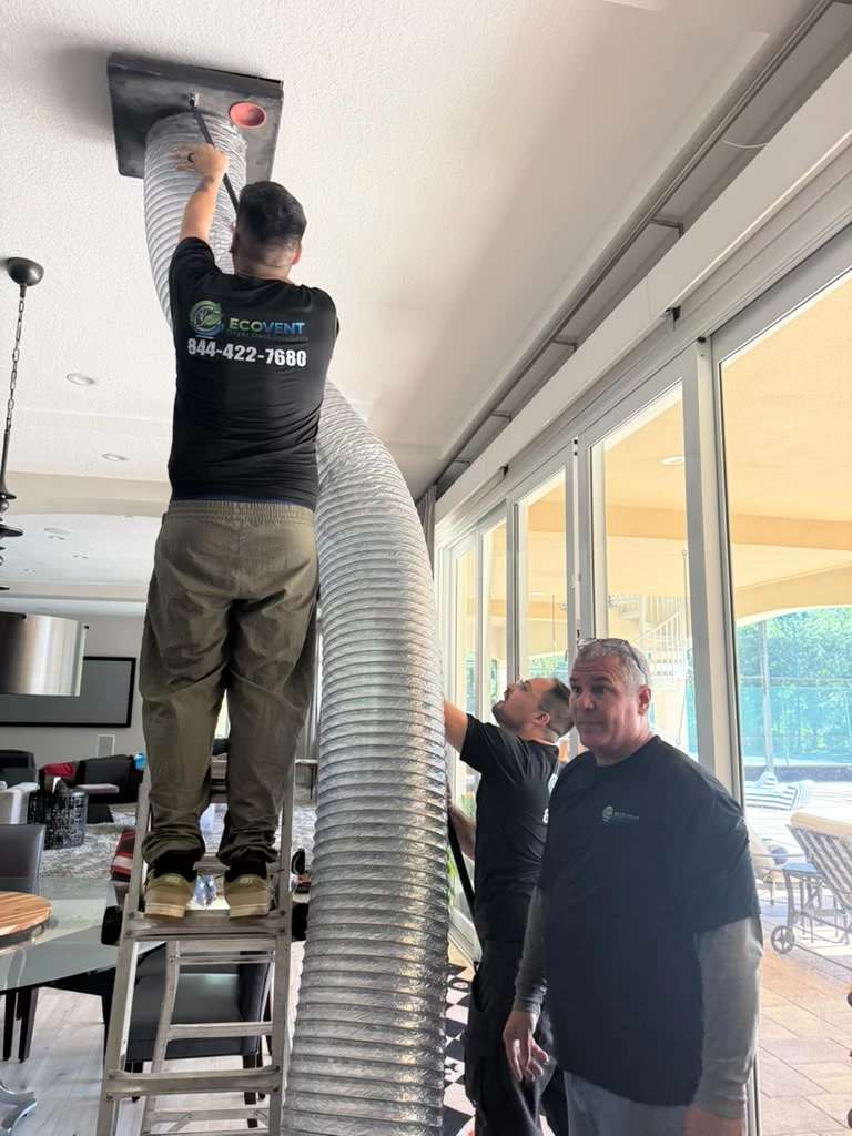 An Ecovent technician on a stepladder feeds a wide flexible duct hose into a ceiling register in a bright living room while two colleagues work below