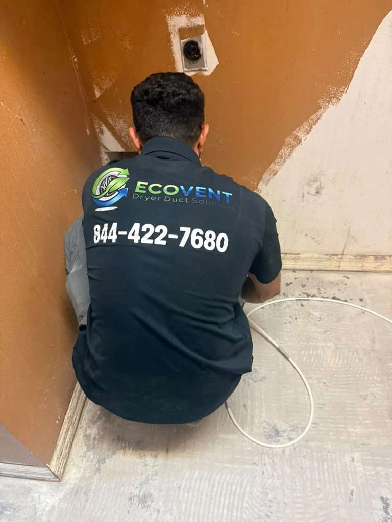 An Ecovent technician in the company shirt, kneeling at a dryer vent connection with the rotary cable coiled beside him