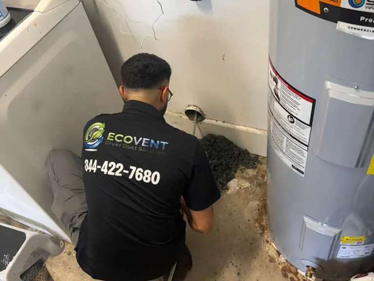 An Ecovent technician clearing a dryer vent at the wall connection in a laundry room, lint pulled out onto the floor