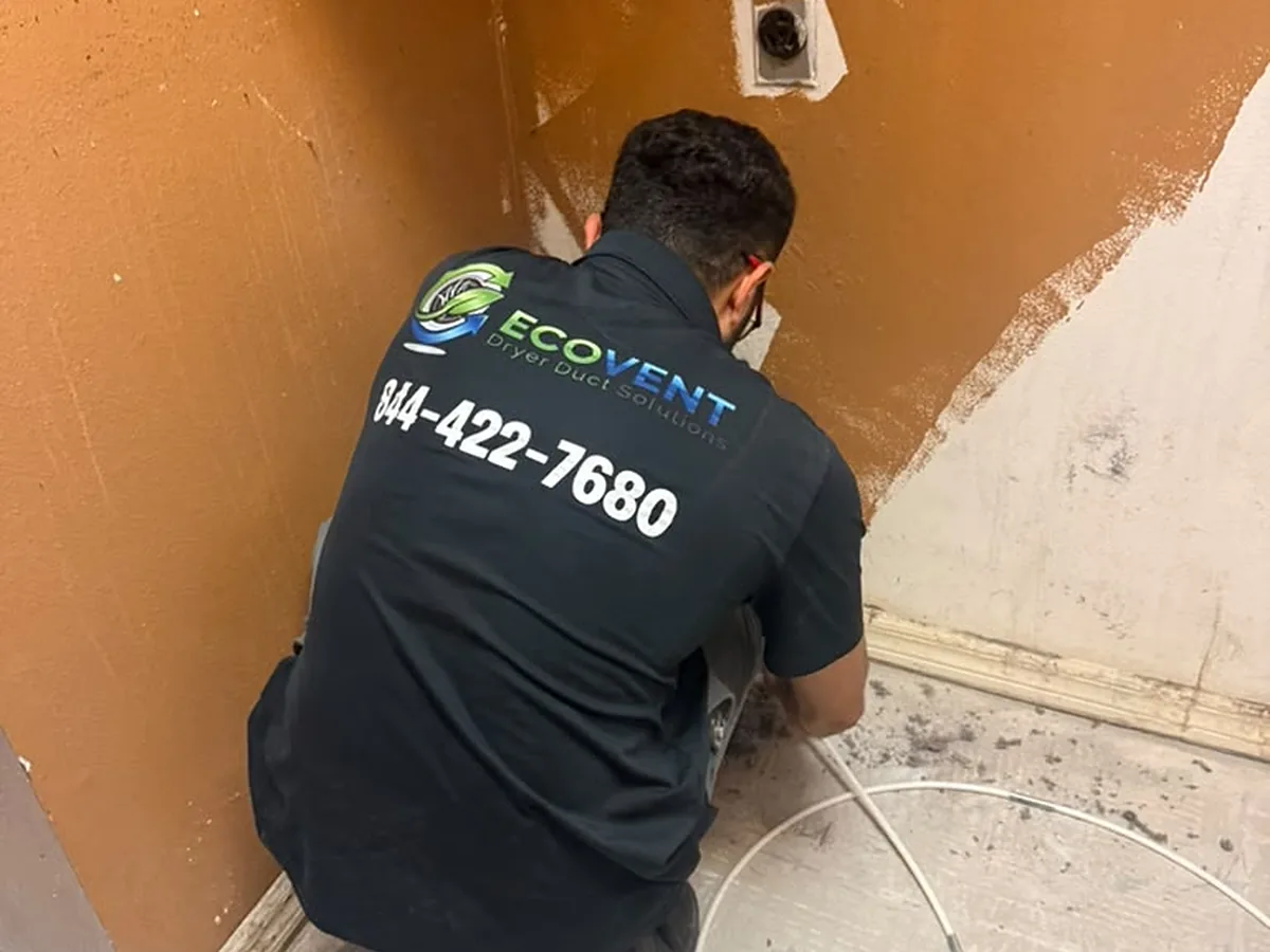 An Ecovent technician kneeling at a dryer vent wall connection, the brush cable feeding into the run and lint on the floor